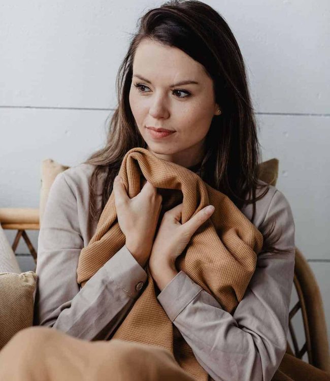 Jonge vrouw met bruin haar op de bank en onder een camelkleurige plaid