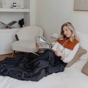 jannekeswarmtesjaals2022 23 vrouw ligt op bed een krant te lezen onder een warme plaid en een warmtesjaal over haar schouders in de kleur cognac