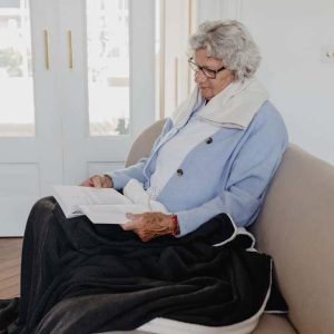 warmtesjaal beige 70 cm 70+ vrouw zit ontspannen op de bank een boek te lezen met over haar benen een grote warme plaid en een warmtesjaal over haar schouders in de kleur beige.